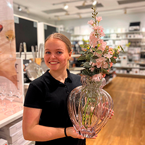 Medarbejder fra Kop & Kande i Slagelse, holder lyserød vase frem i butik. Der er lyserøde blomster i vasen også.